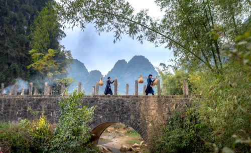 Free Two people in traditional attire cross a stone bridge in a lush, mountainous landscape. Stock Photo