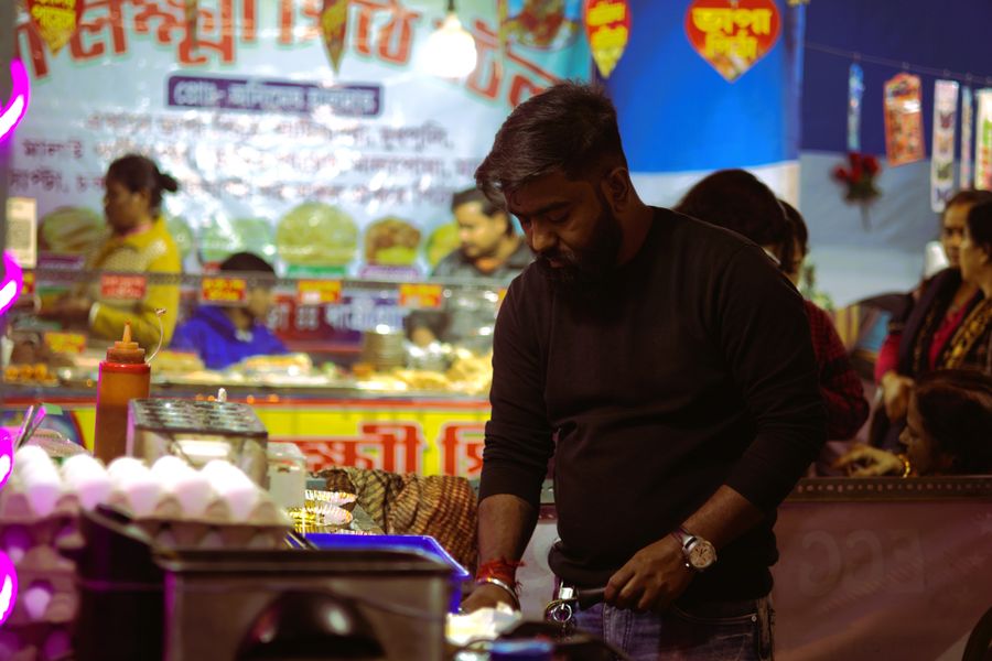 Street food vendor cooking at a vibrant night market with colorful signs and crowd in the background.