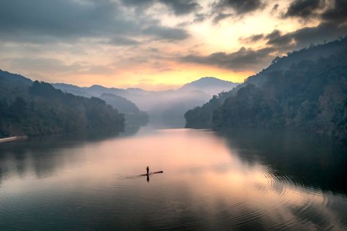 Free A lone fisherman paddles across a tranquil mountain lake at sunset, surrounded by misty hills. Stock Photo