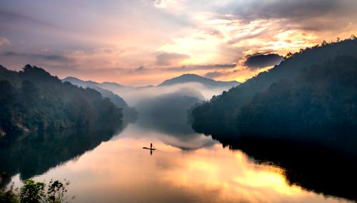 Free A lone figure paddles on a tranquil lake surrounded by misty mountains at sunset. Stock Photo