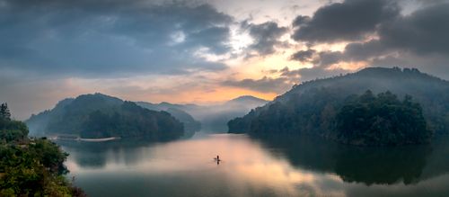 Free Tranquil morning scene with a solitary boat on a misty lake surrounded by forested hills. Stock Photo