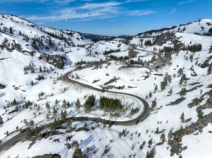 Aerial view of snow-covered Donner Pass with a winding mountain road and clear blue skies.