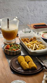A delicious homemade meal featuring fresh corn, fries, salad, and juice on a rustic table.