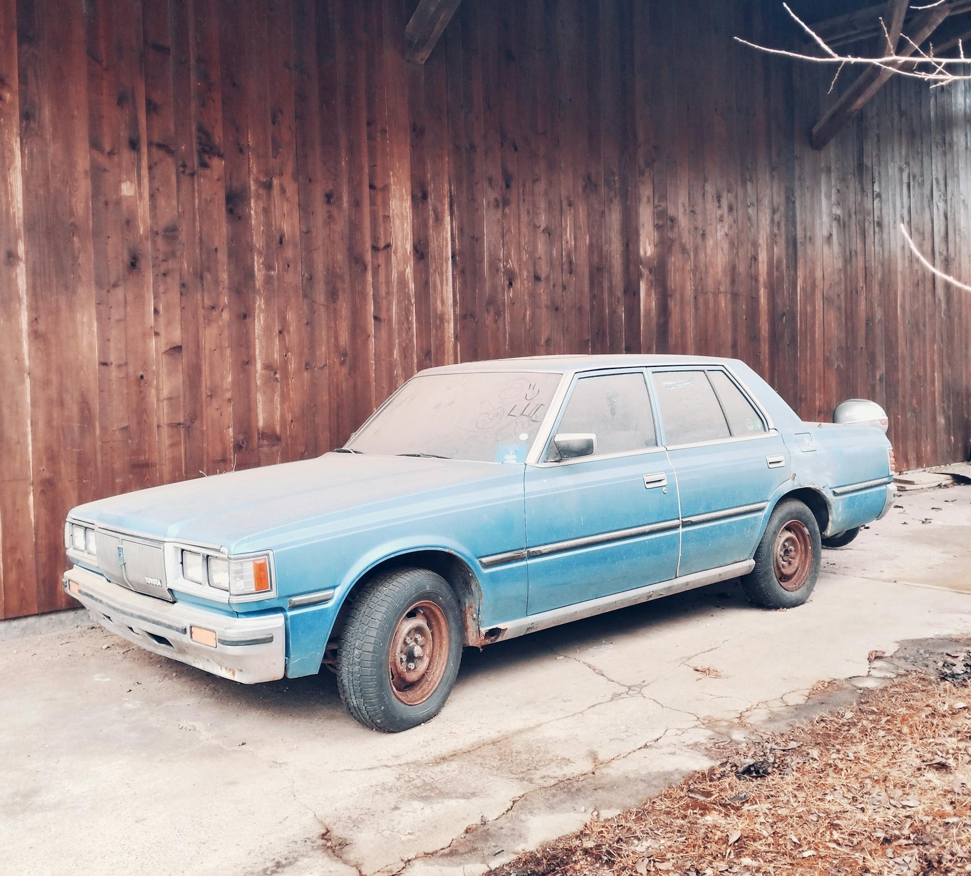 Dusty old blue car parked against a rustic wooden wall, showcasing vintage charm.