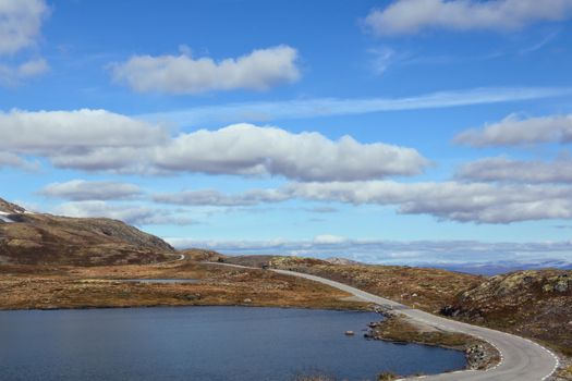 Winding road along a serene lake in the Norwegian mountains under a bright blue sky.