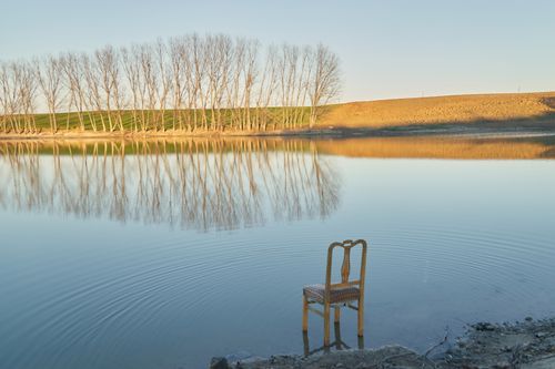 Free A lonely chair stands submerged in a calm lake reflecting bare trees at sunset. Stock Photo