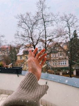 A person's hand in a sweater reaching towards a snowy outdoor scene with houses and bare trees.