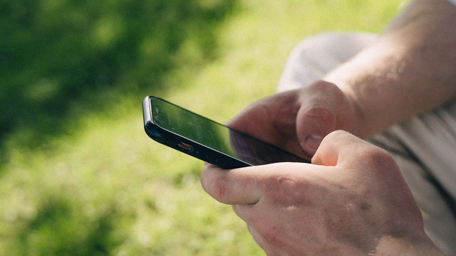 Hands holding smartphone outdoors in a sunny park. Ideal for tech and lifestyle themes.