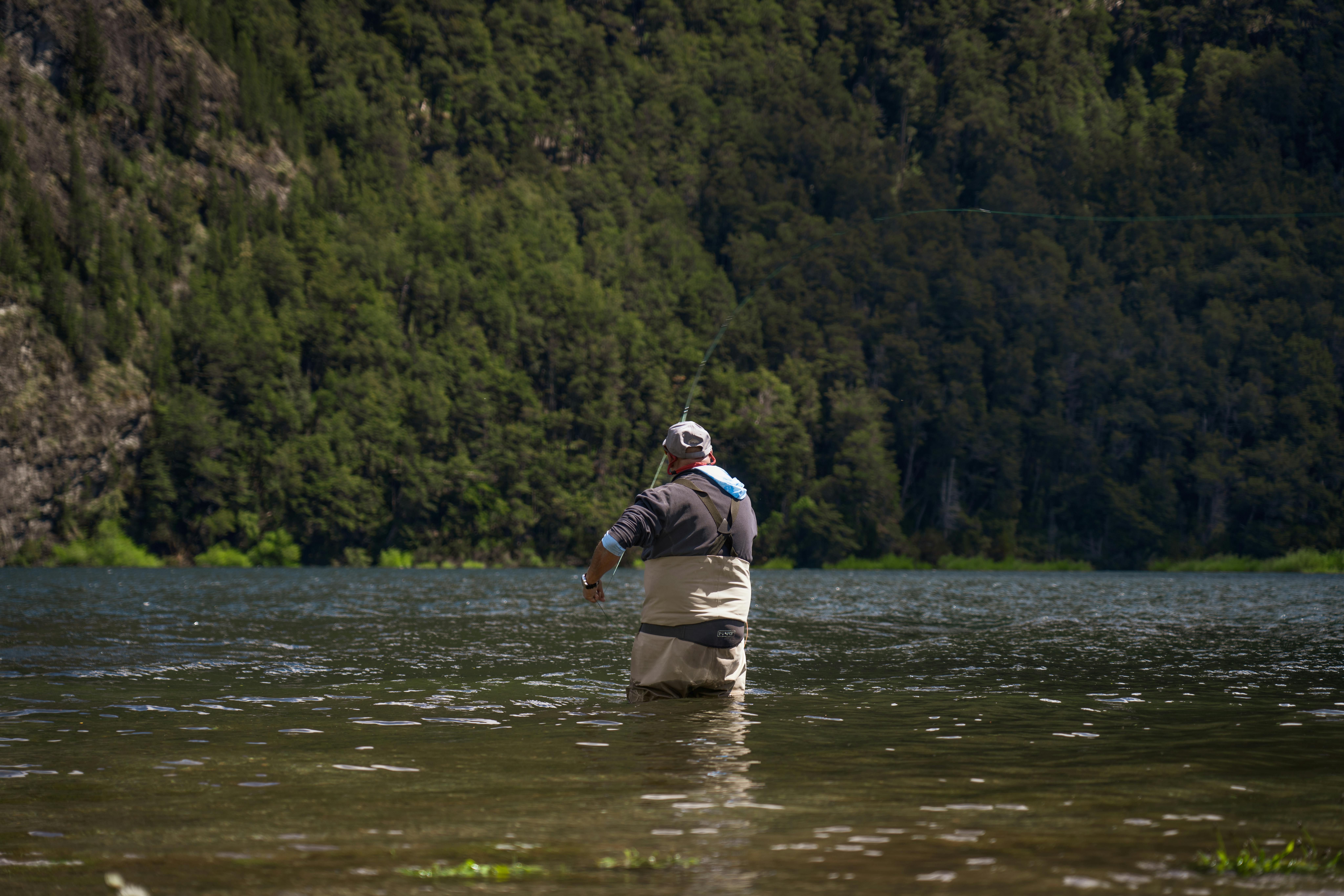 Man fly fishing in serene Patagonian lake surrounded by lush, green mountains.