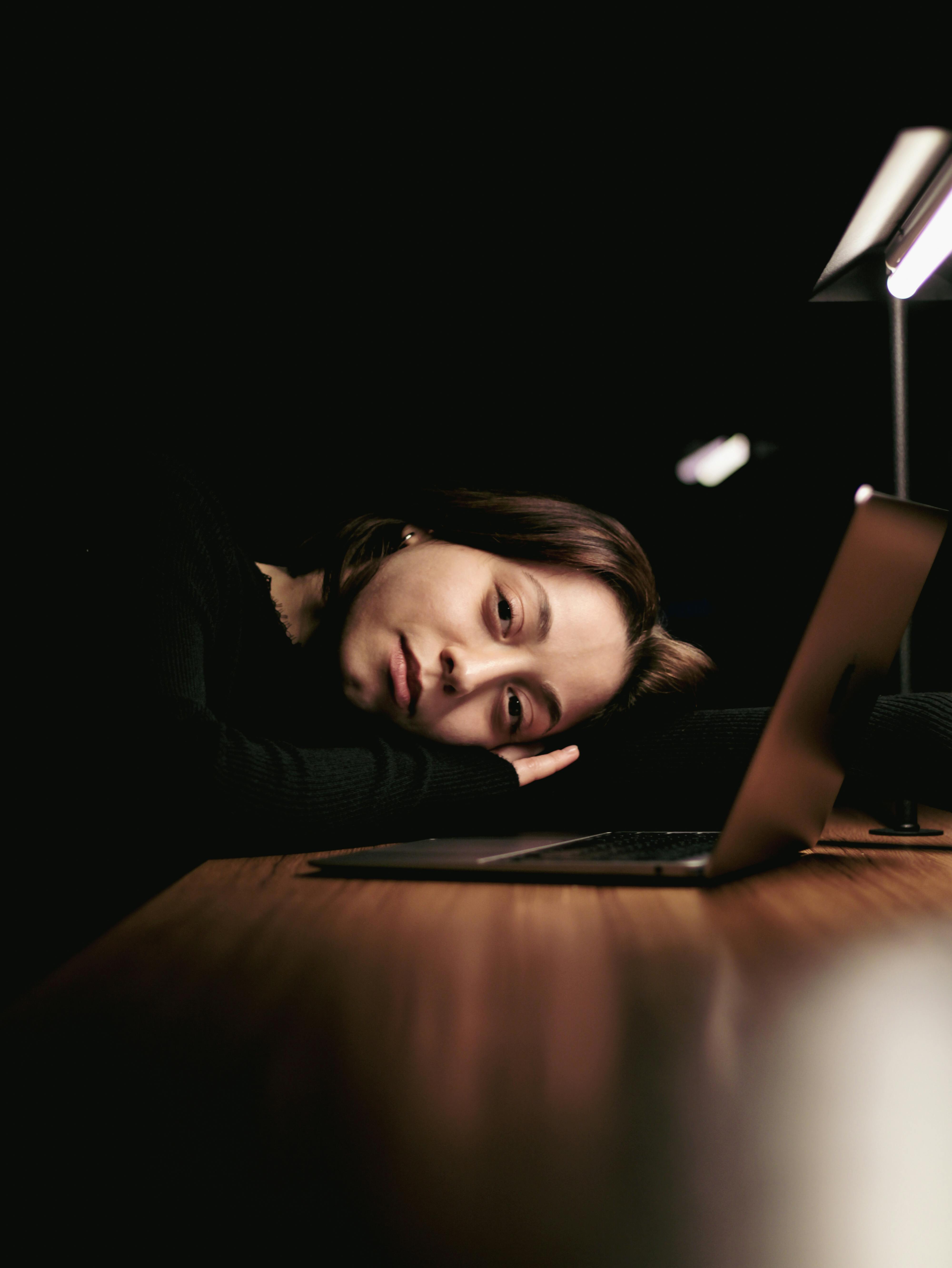 woman resting head on desk with laptop and lamp