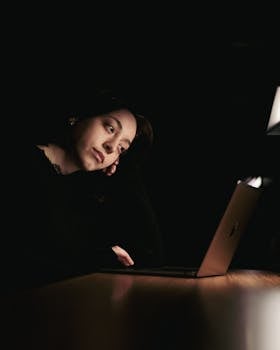 A thoughtful woman in dim lighting works on her laptop late at night, reflecting focus.