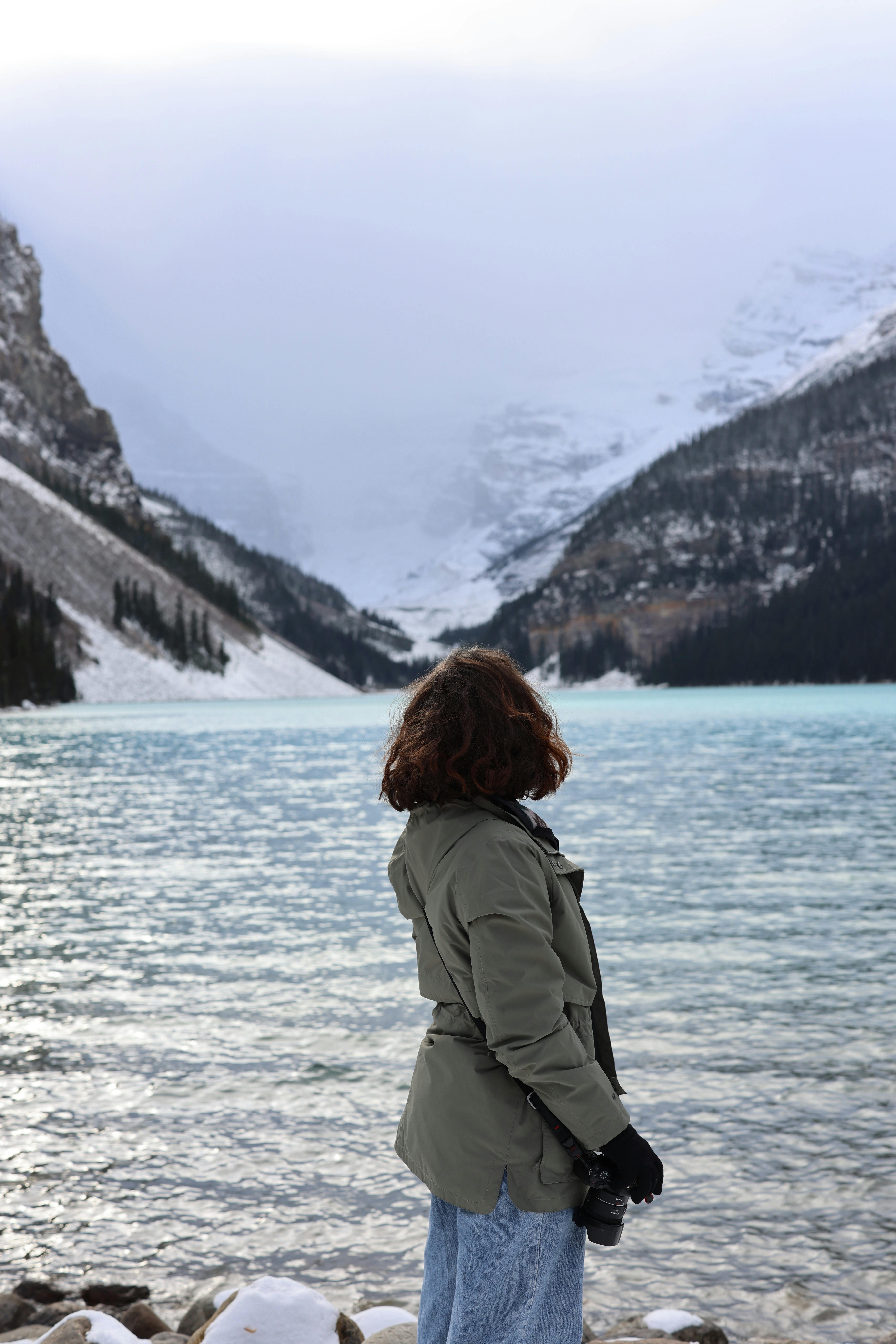 Woman in winter clothing stands by a tranquil glacier lake surrounded by snow-capped mountains.