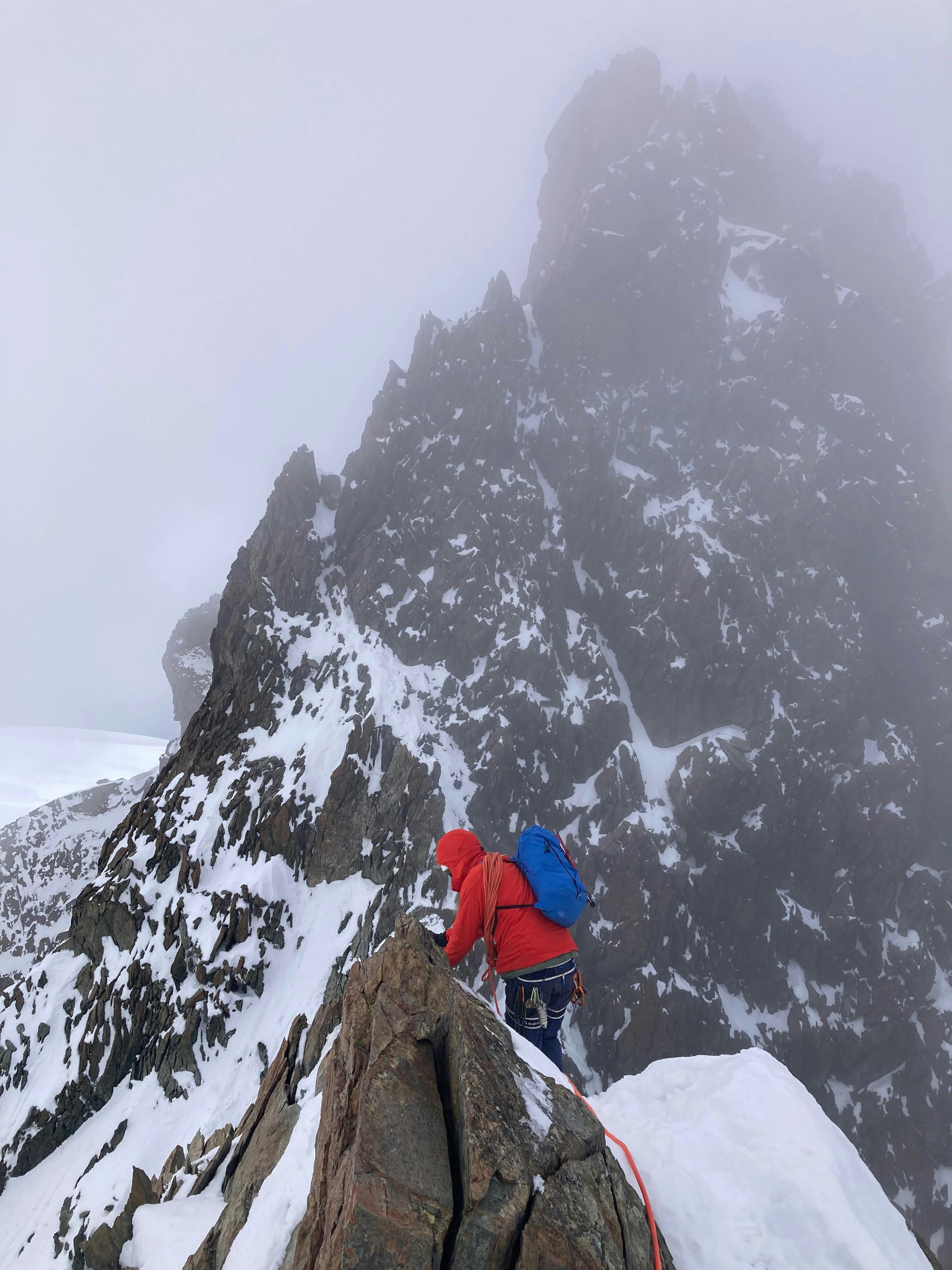 Mountaineer in red jacket scaling a rugged snow-covered ridge with ice axe
