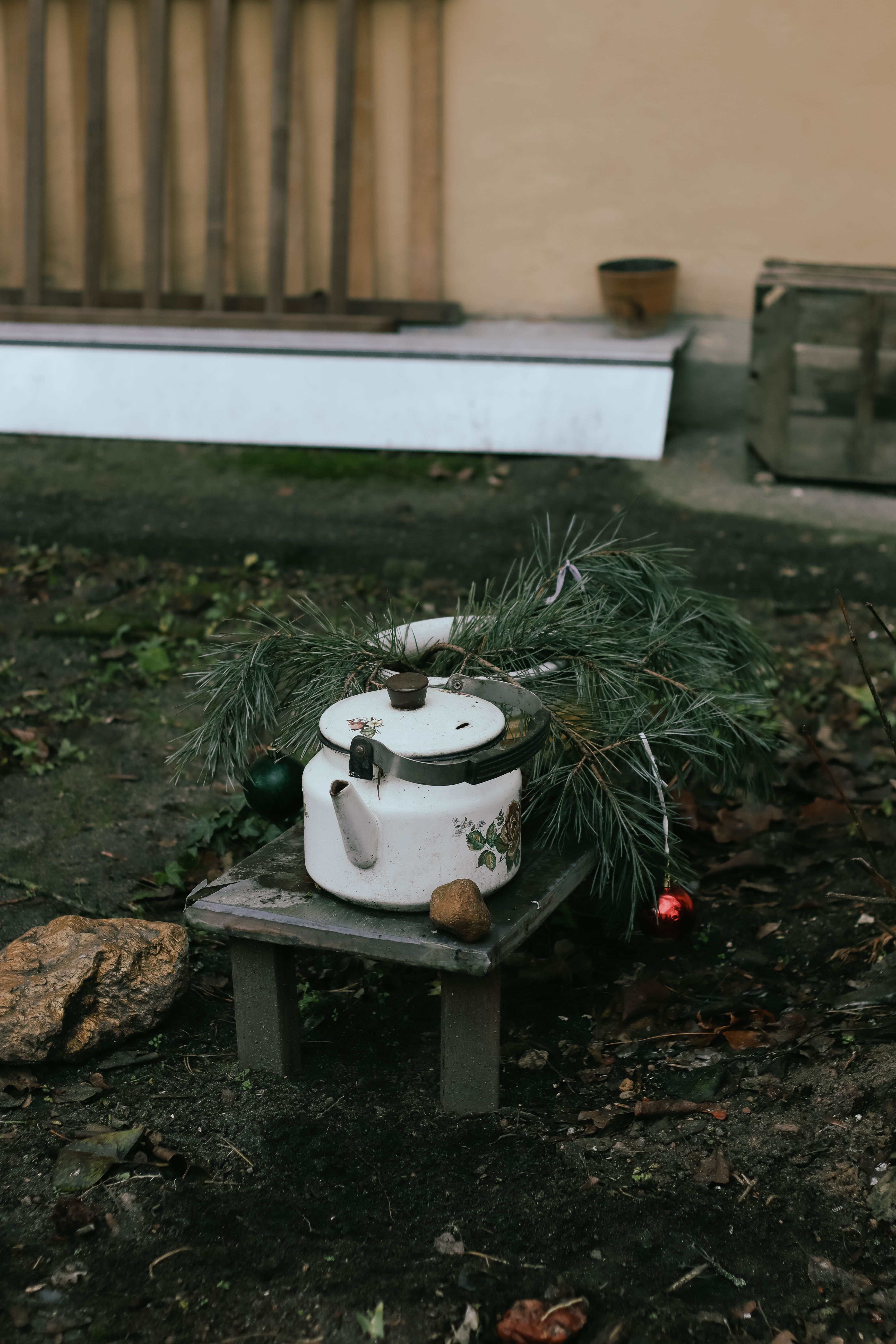 A white vintage kettle on a small wooden table outdoors, decorated with pine branches.