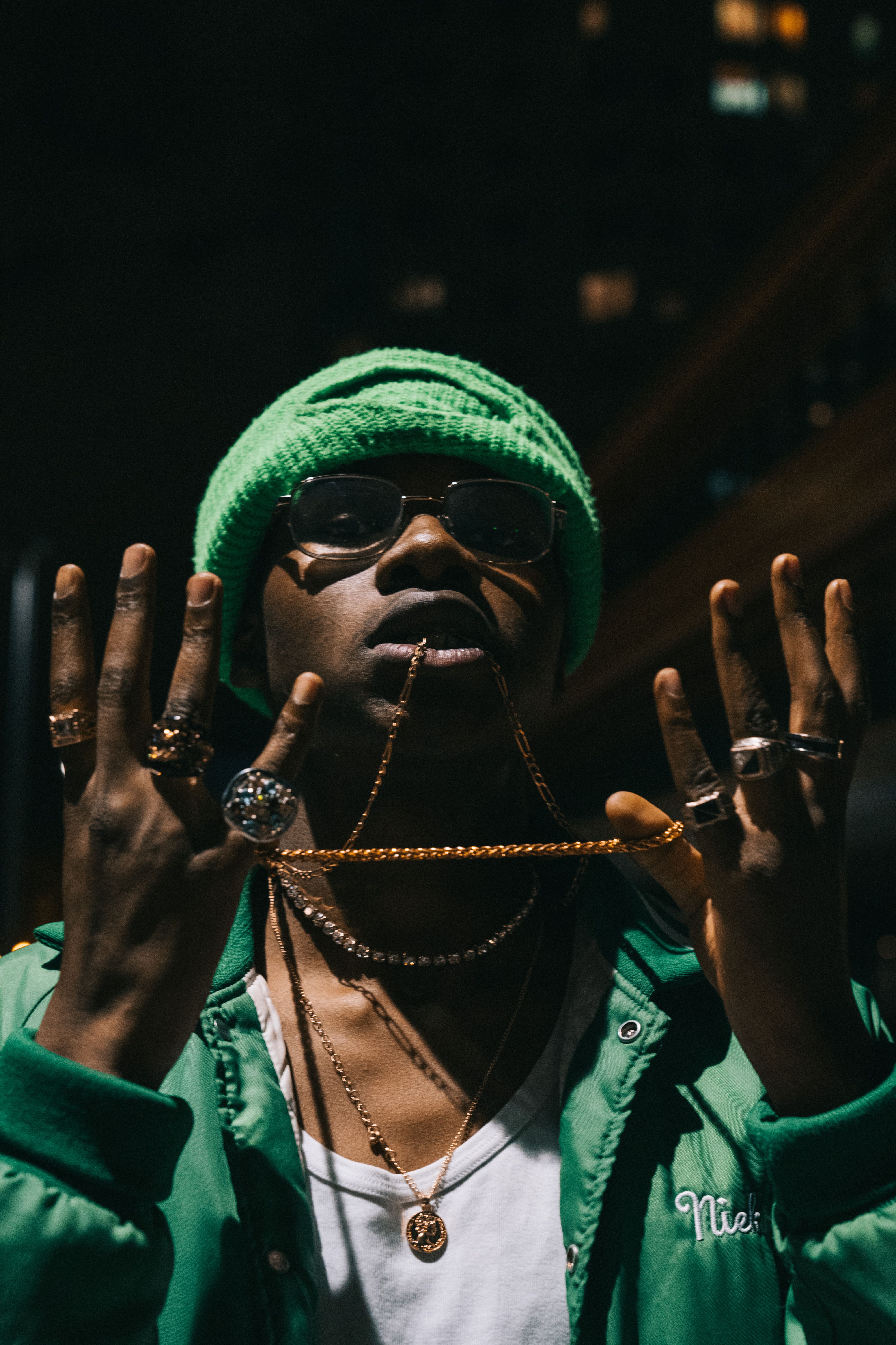 Young man in green jacket and knit cap showcasing jewelry at night with urban backdrop.
