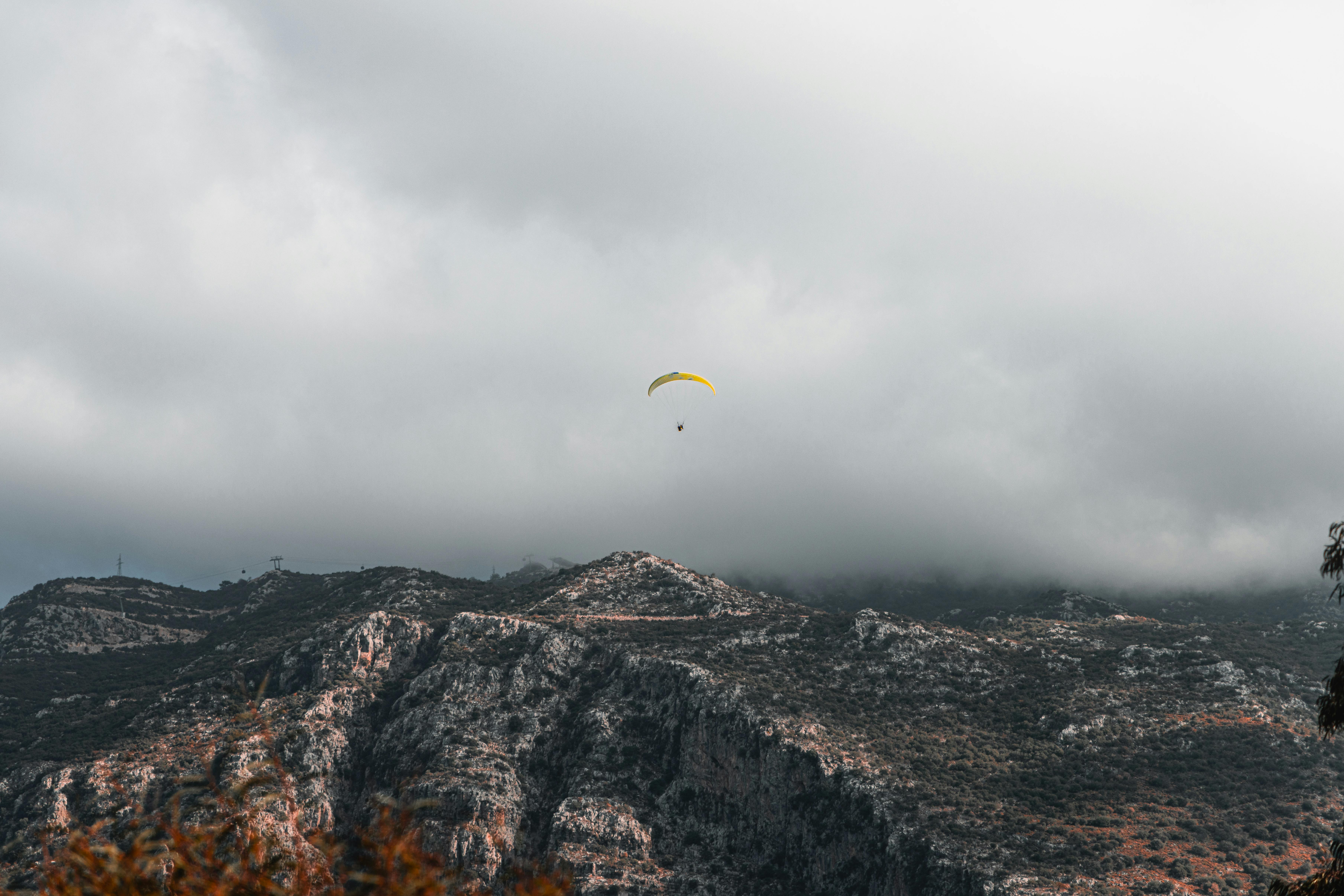A paraglider gracefully flies over rugged mountainous terrain beneath a cloudy sky, showcasing adven