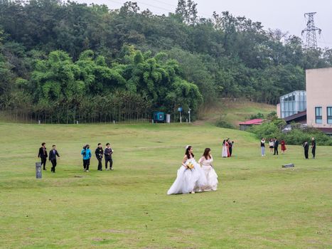 Two brides in wedding dresses walk on a grassy field with guests in the background.