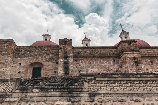Ancient Zapotec architecture with intricate stonework and iconic red domes under a cloudy sky.