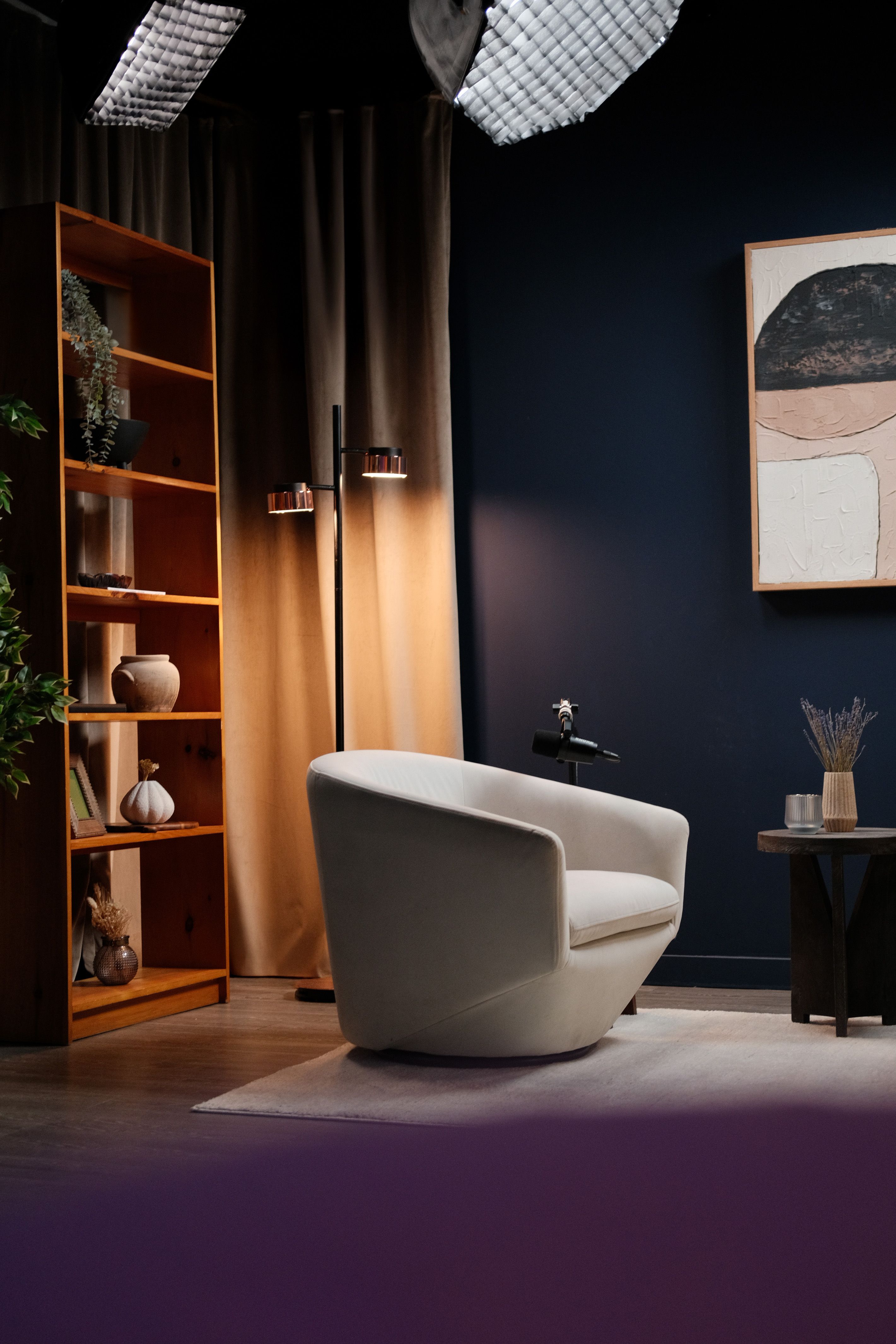 Podcast set with a white armchair, a wooden bookshelf, and warm lamp light against a navy wall