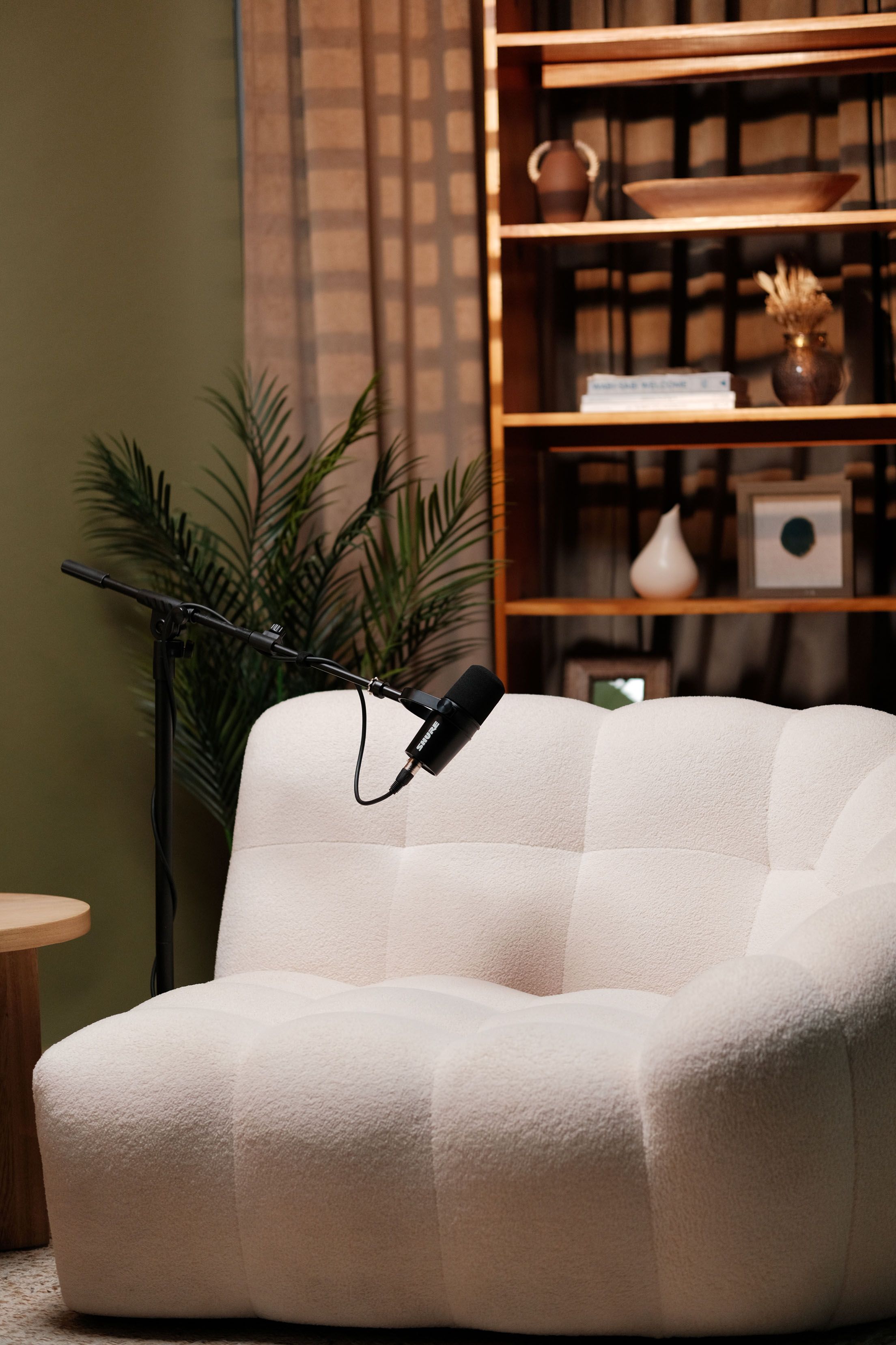 A microphone stand beside a cream sofa and a wooden bookshelf on a green podcast set