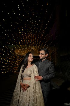 Charming couple in traditional attire posing under a canopy of twinkling lights.
