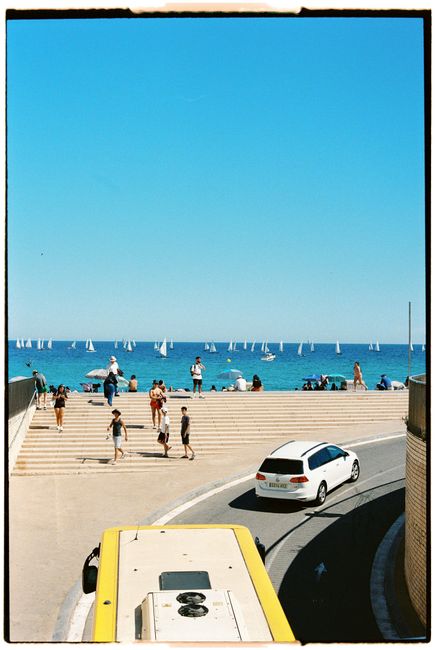 Vibrant beach scene with sailboats, people, cars, and a clear blue sky.