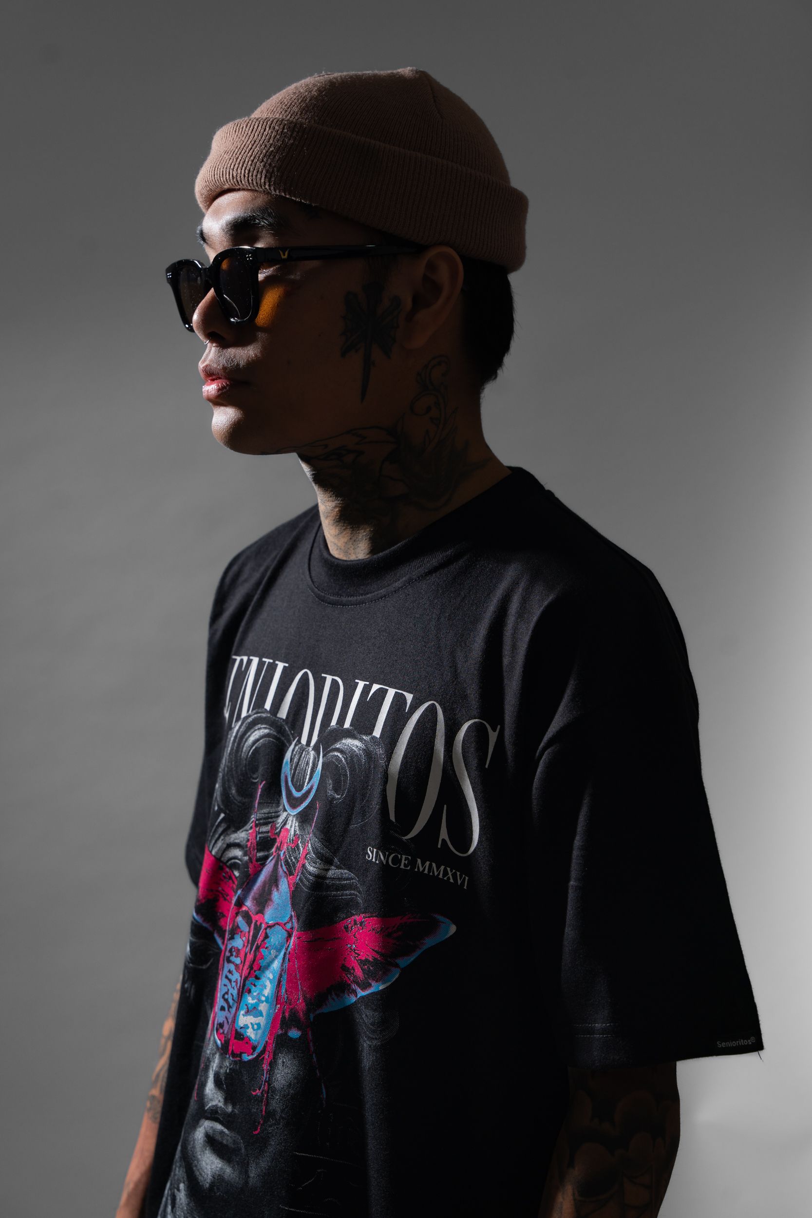 A trendy tattooed man in sunglasses and graphic tee poses in a studio setting.