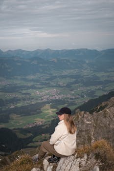 A woman in casual attire sits atop a mountain peak, enjoying the view.