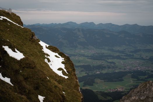 Dramatic mountain landscape with snow patches on a cliff, overlooking a lush valley.