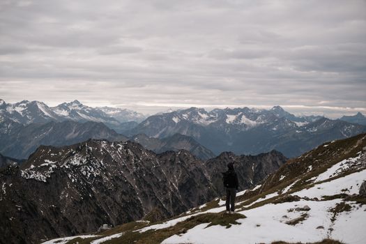Solitary hiker observing snow-capped mountain range under overcast skies, capturing serene adventure.