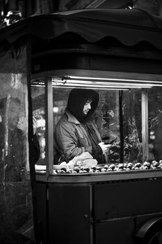 Black and white photo of a street vendor selling chestnuts from a cart at night.