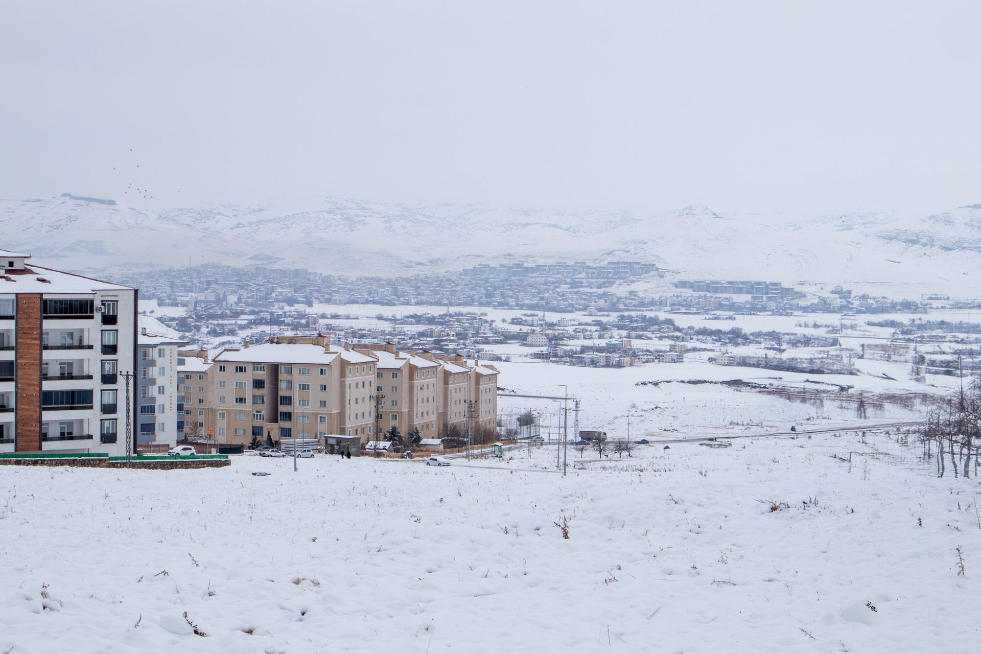 Snow-covered landscape with apartment buildings and distant city view in the background.