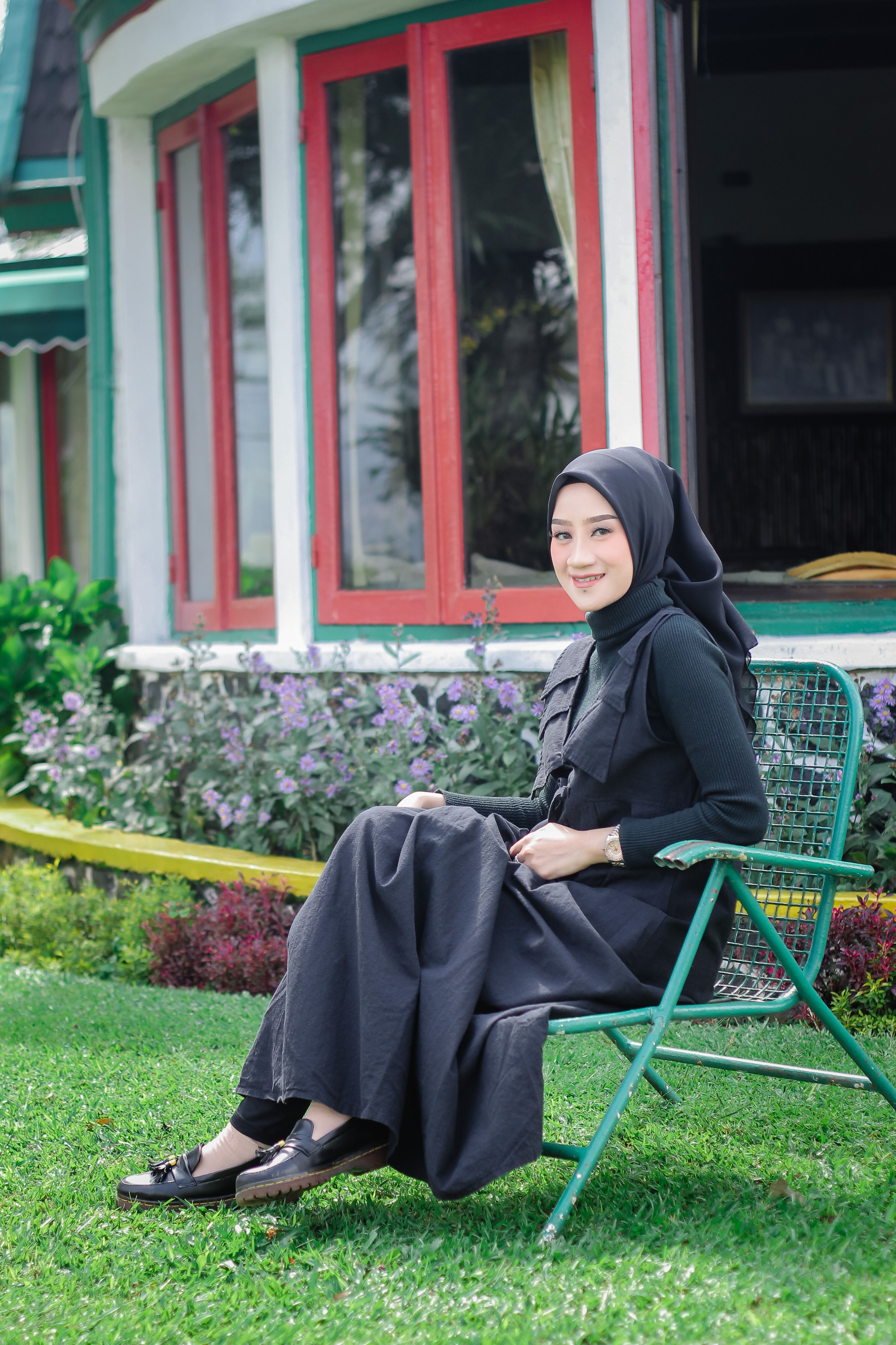 Woman in black hijab sitting on a chair in a garden, smiling warmly.