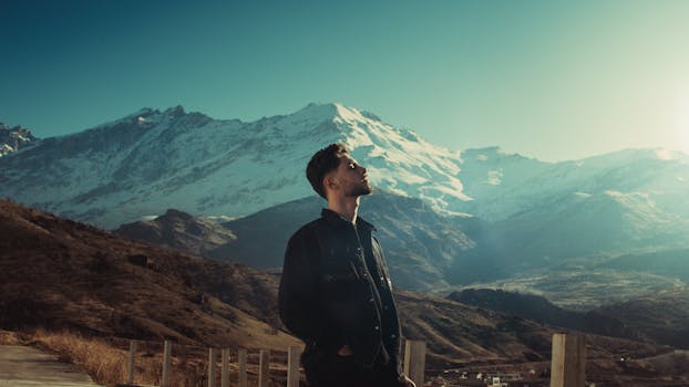 A man stands thoughtfully with snowy mountains in Kurdistan, Iran in the background, under a clear blue sky.