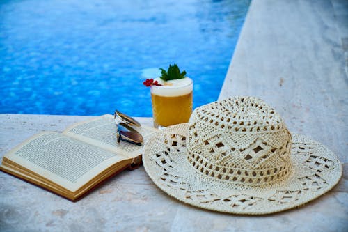 Beige Straw Hat, Book, Sunglasses, and Drink Beside Pool