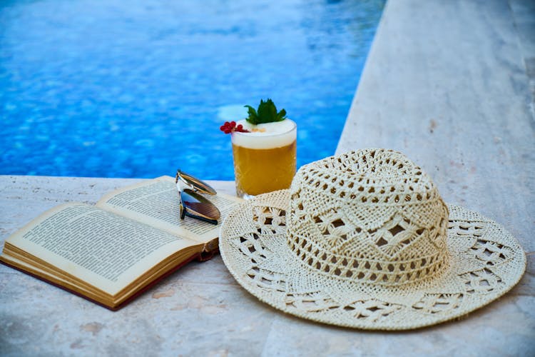 Beige Straw Hat, Book, Sunglasses, And Drink Beside Pool