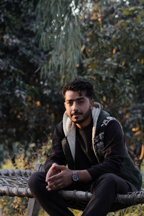 Free A young man in a hoodie sits on a woven bench amidst lush greenery, enjoying a peaceful moment outdoors. Stock Photo