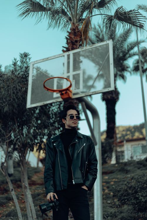 Free Fashionable man with camera poses stylishly by an outdoor basketball hoop. Stock Photo