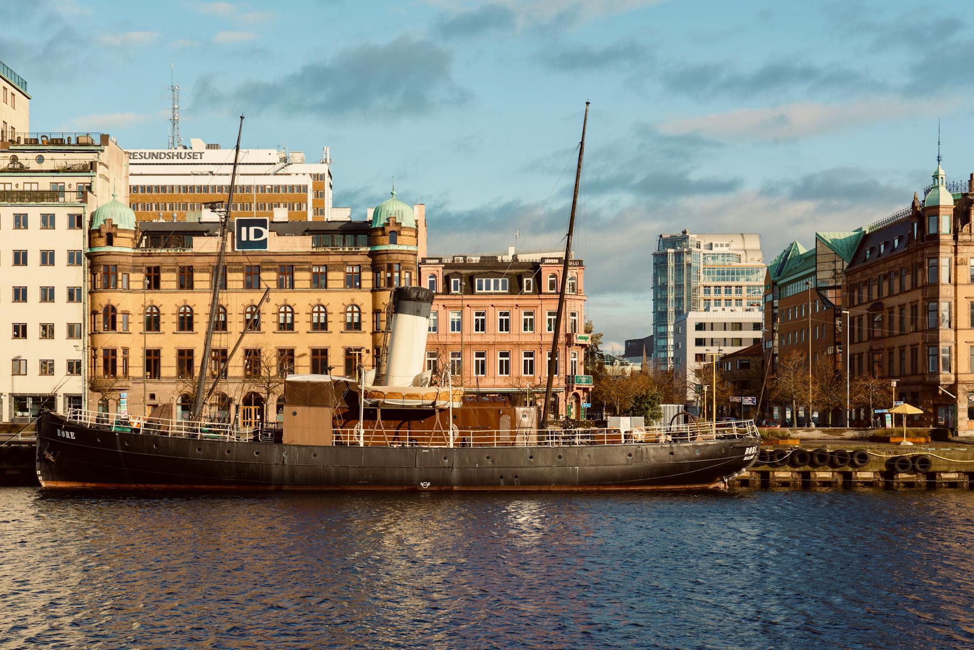 Photograph of a historic ship docked in a bustling riverfront cityscape under a bright blue sky.