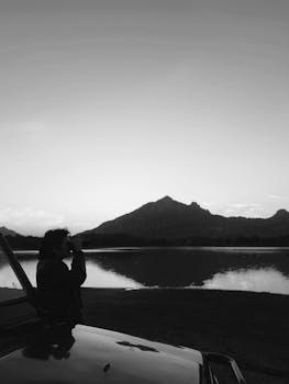 A person using binoculars near a mountain and lake during twilight.