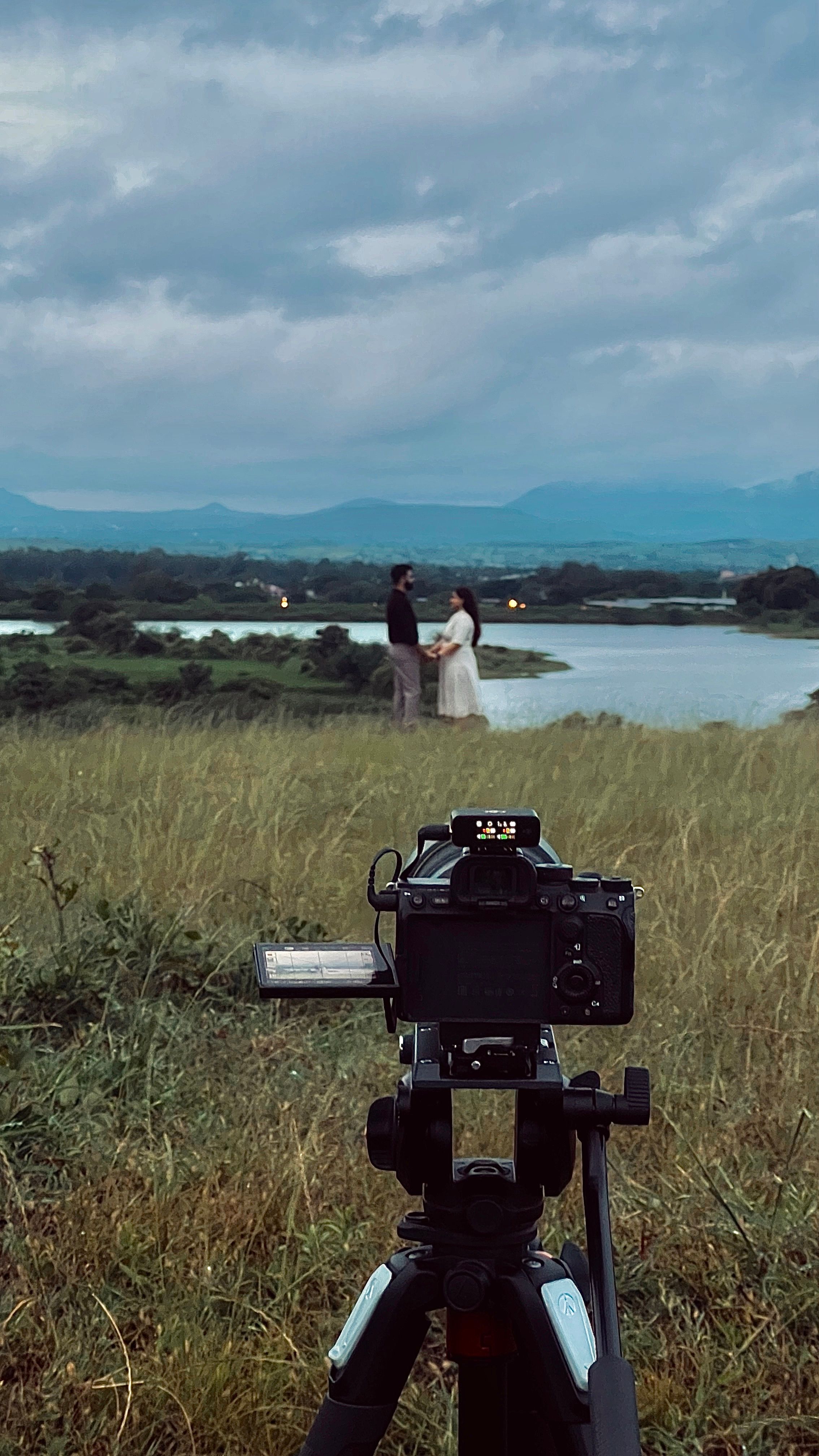A couple stands by a river in a scenic outdoor setting, captured through a camera on a tripod.