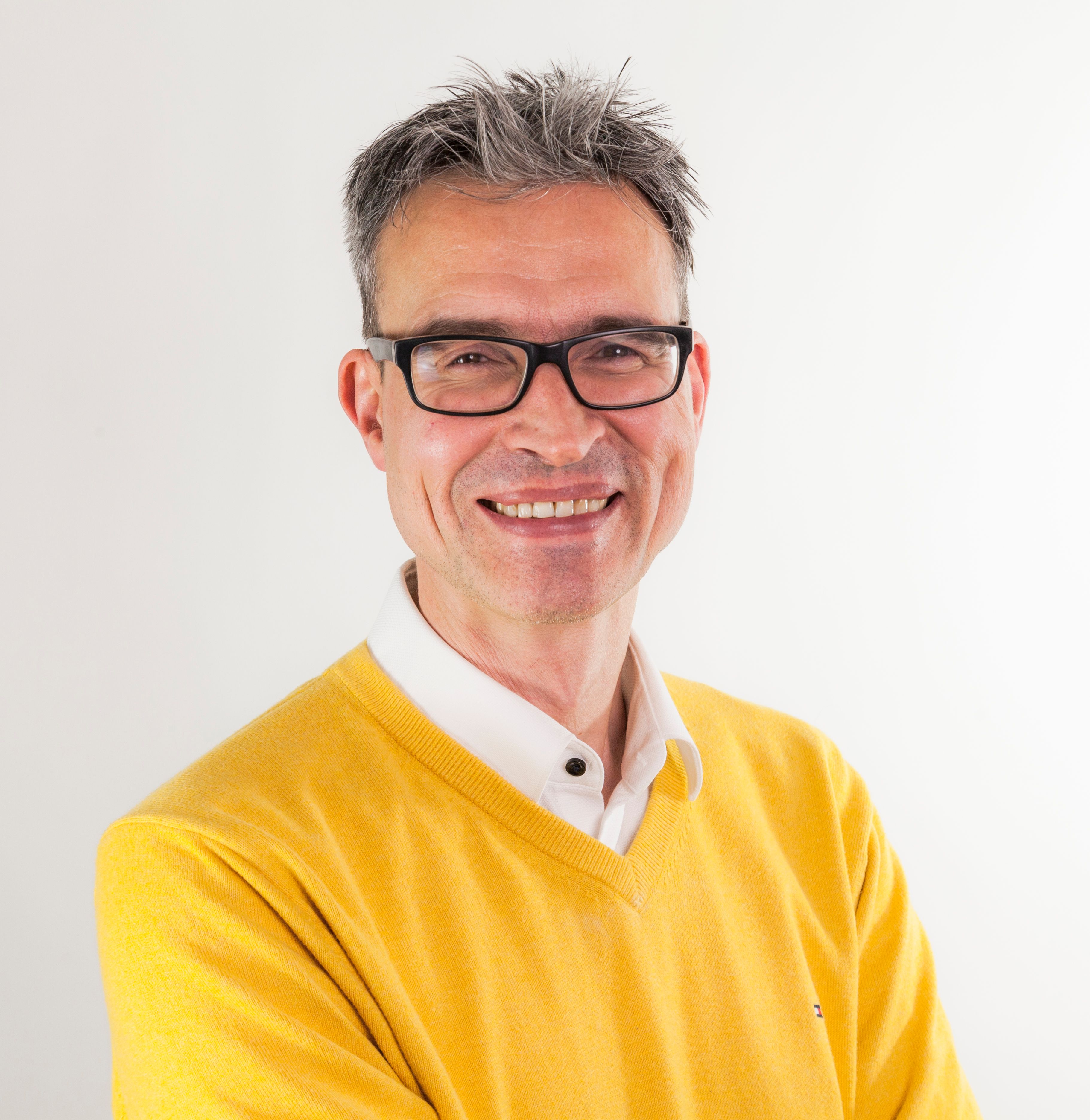 Confident adult man wearing glasses and yellow sweater, smiling at camera.