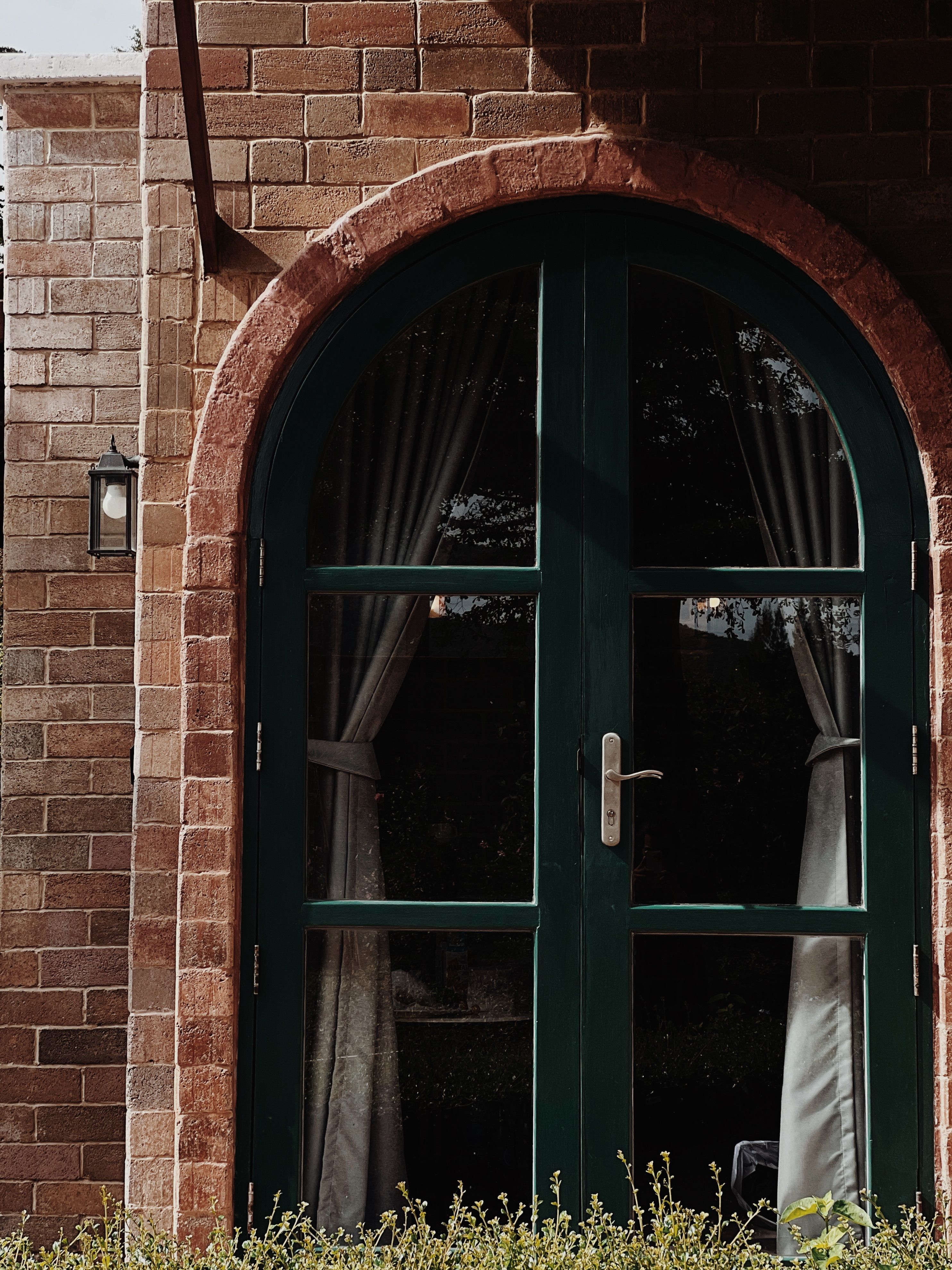 A sunlit arched doorway with green trim and curtains framed by red brick wall.