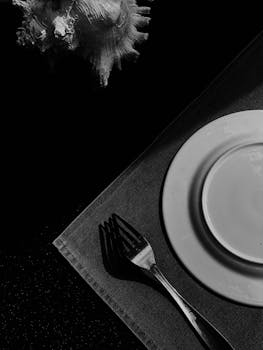 Black and white artistic photograph of a table setting with plates, fork, and seashell.