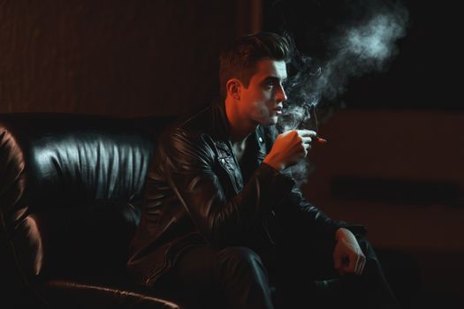 在光线昏暗的房间里，一名穿着皮夹克的男子坐在沙发上抽烟