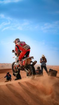 Motorcyclist racing through desert sand dunes with photographers capturing the action.
