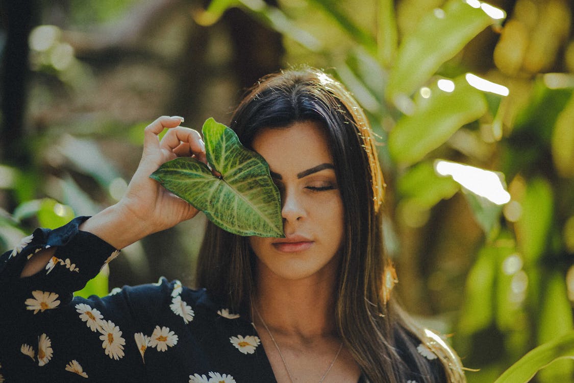 Selective Focus Photo of Woman in Black Floral Top Posing With Her Eyes Closed While Holding Green Leaf Over Right Eye