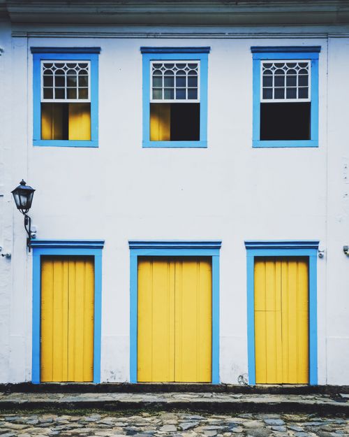Free Charming colonial building facade featuring vibrant yellow doors and blue window frames against a white wall. Stock Photo