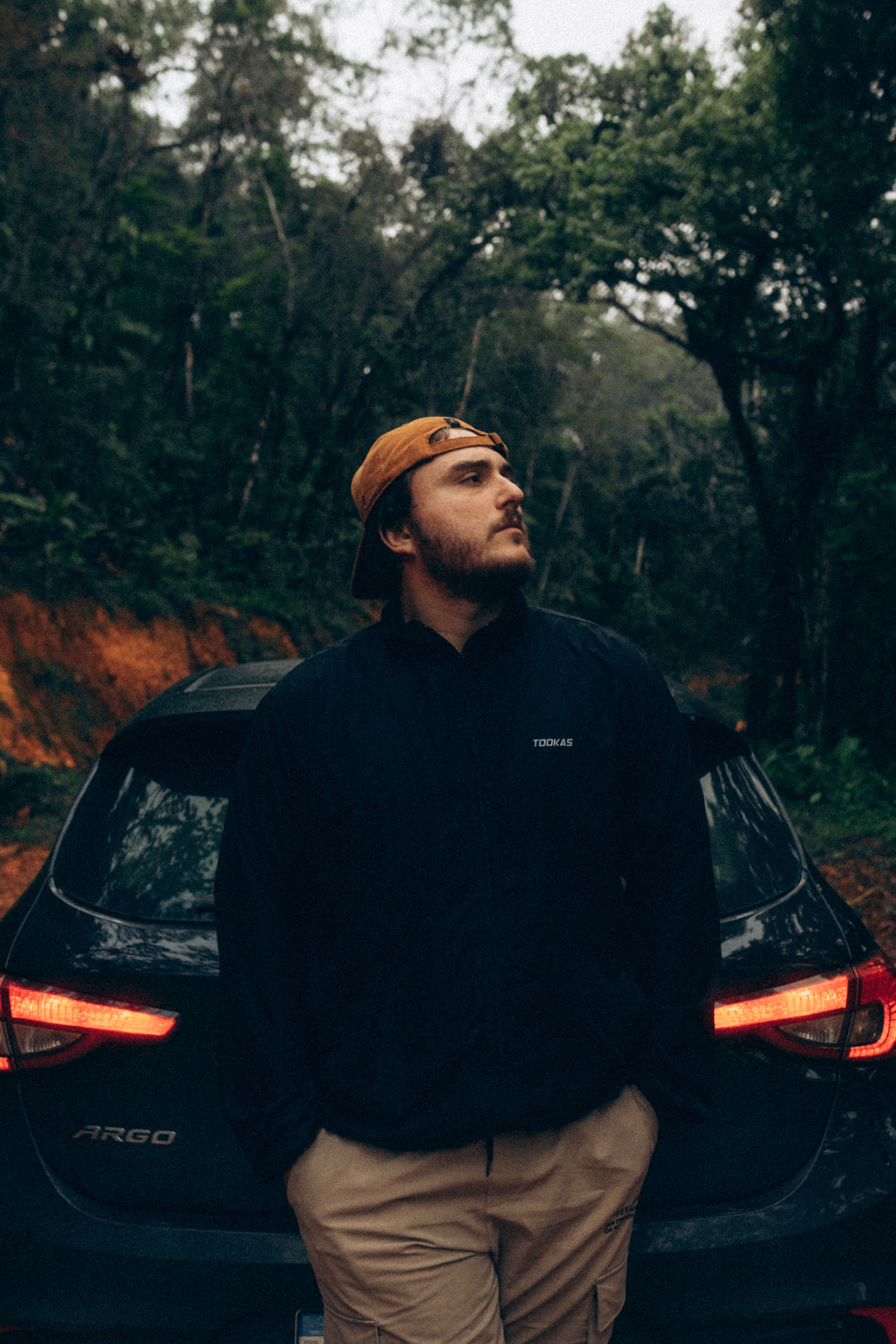 A young man stands confidently by a car in a lush forest landscape, embodying adventure.
