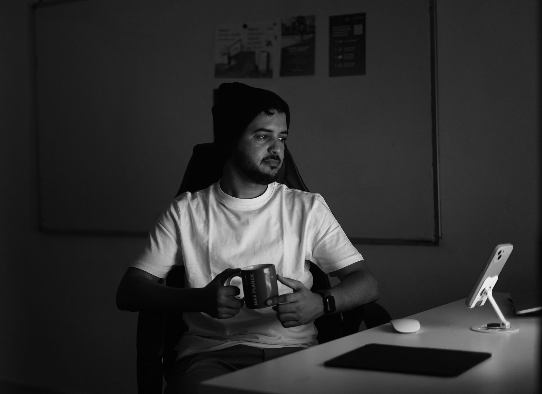 Black and white photo of a man holding a mug in a dimly lit office, deep in thought.